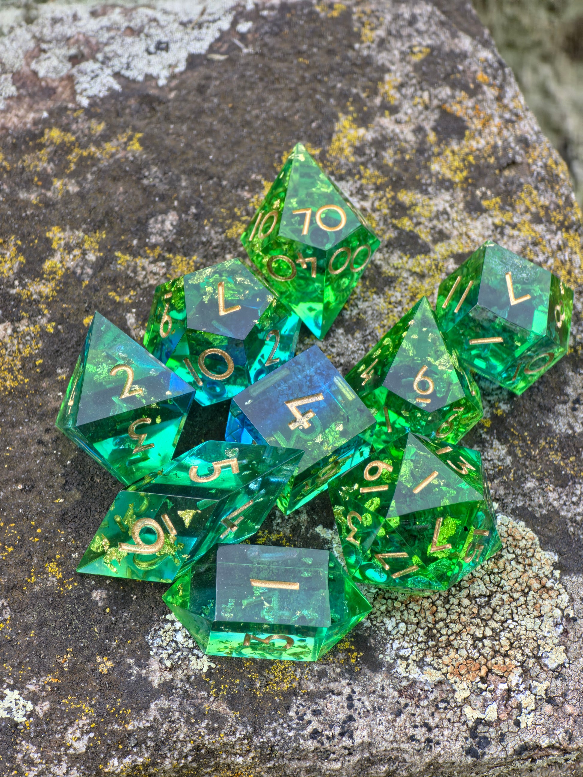 Komplettes „Emerald Tide“ Resin-Würfelset mit goldenen Flakes, glänzend im natürlichen Licht auf Stein arrangiert.
Complete “Emerald Tide” resin dice set with gold flakes, shining in natural light and arranged on stone.