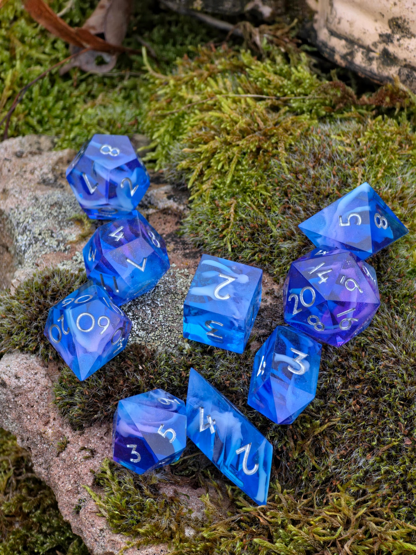 Polyedrische DnD-Würfel in Blautönen mit lila Schimmer, dekorativ auf Moos arrangiert.
