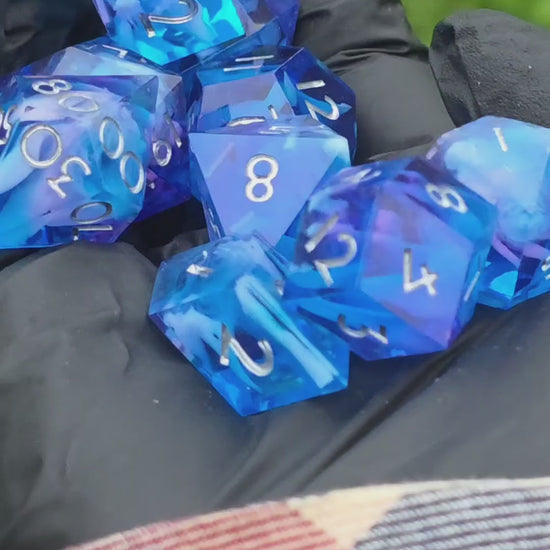 Kurzes Video eines handgemachten Cloud Nebula Dice Sets in Blau mit sanftem lila Schimmer, das im Licht glitzert. Zeigt Nahaufnahmen der Würfel und ihre Bewegung.

