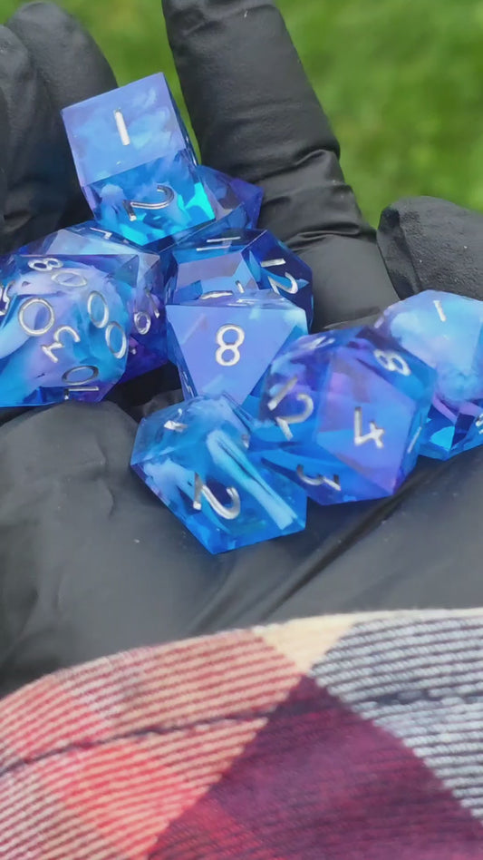Kurzes Video eines handgemachten Cloud Nebula Dice Sets in Blau mit sanftem lila Schimmer, das im Licht glitzert. Zeigt Nahaufnahmen der Würfel und ihre Bewegung.
