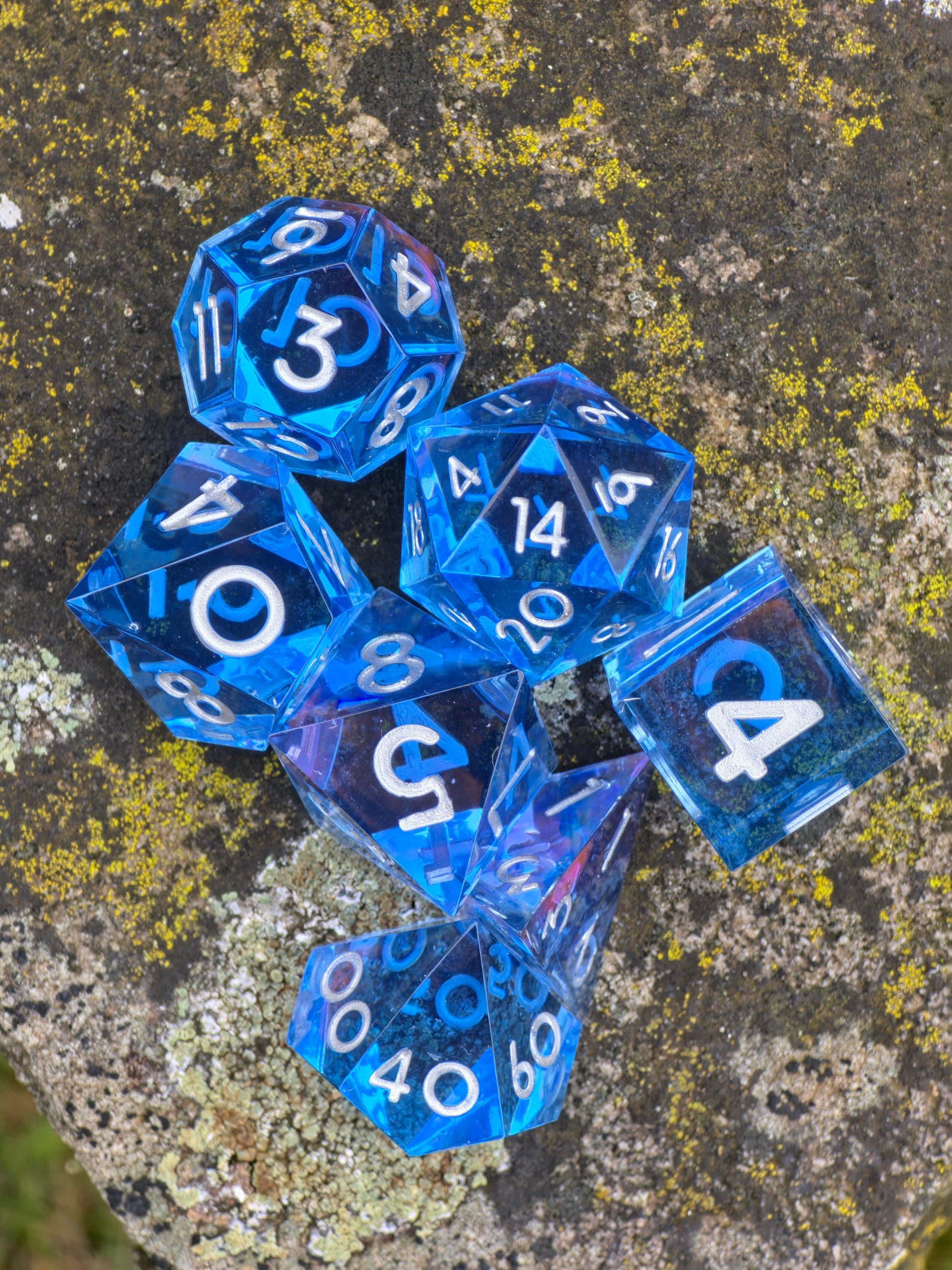 Detailaufnahme des „Abyss of the Sapphire Tide“ Würfelsets, transparente blaue Resinwürfel mit klaren silbernen Zahlen.  
Close-up of the “Abyss of the Sapphire Tide” dice set, transparent blue resin dice with crisp silver numbers.