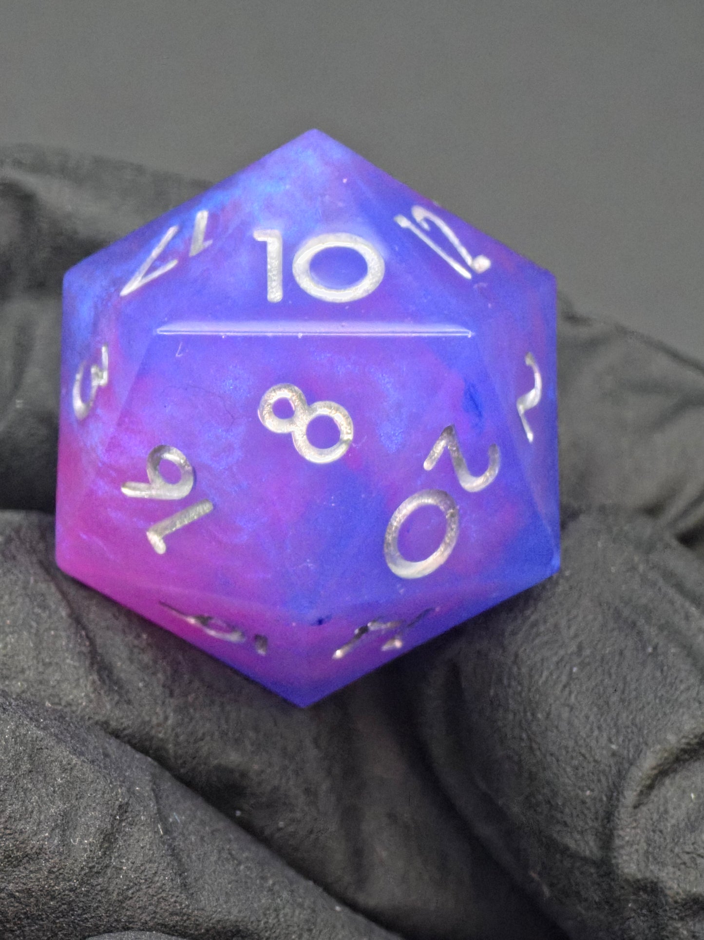Handaufnahme des „Aetherfall D20“, lila-blauer Resinwürfel mit silbernen Zahlen, Standardgröße. 
Hand shot of the “Aetherfall D20”, purple-blue resin die with silver numbers, standard size.