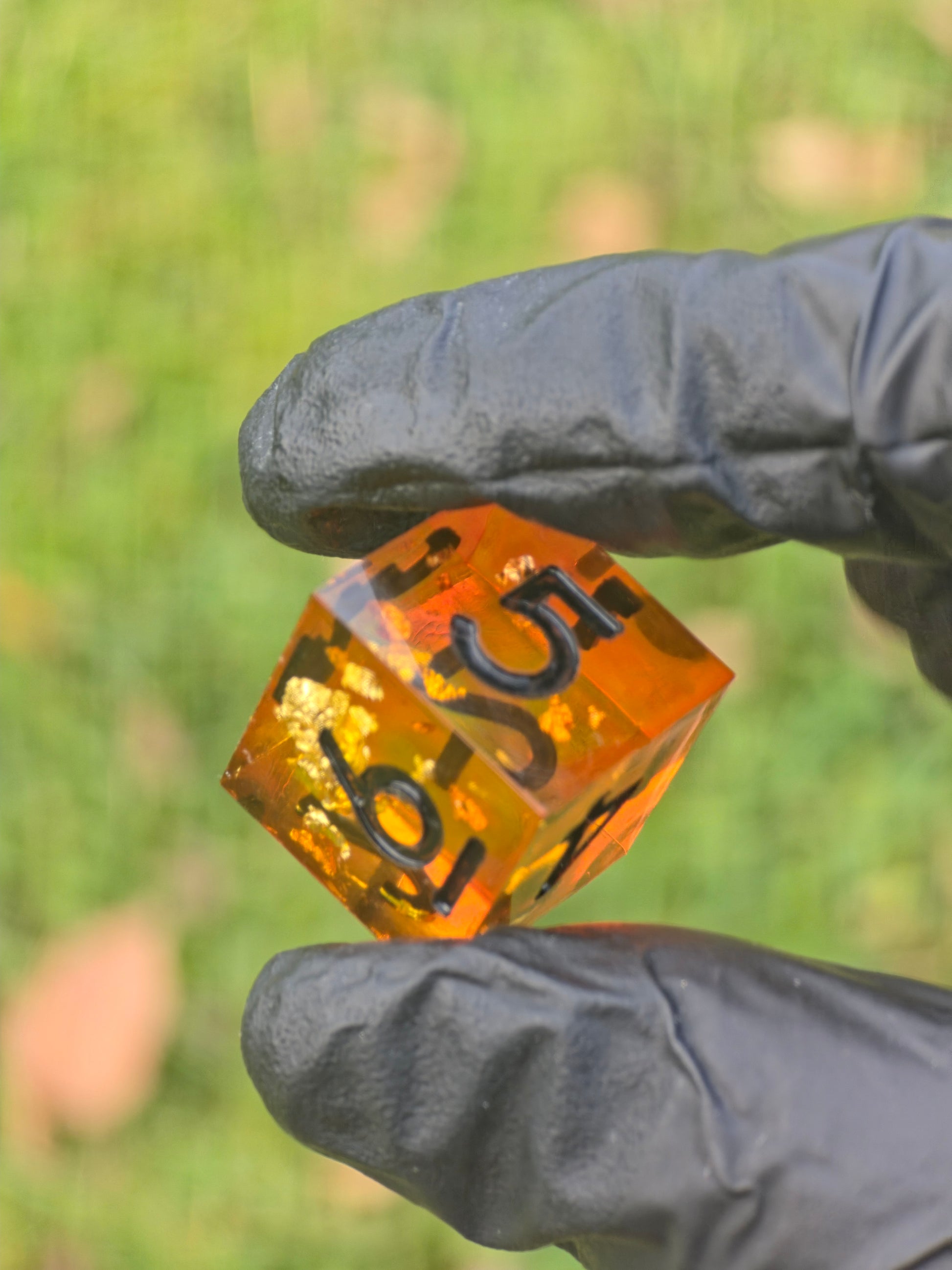 Einzelner D6 „Bernstein Fragment“ – amber-orangefarbener Resinwürfel mit goldenen Partikeln in der Hand.  
Single D6 “Amber Fragment” – amber-orange resin die with gold flakes held in hand.