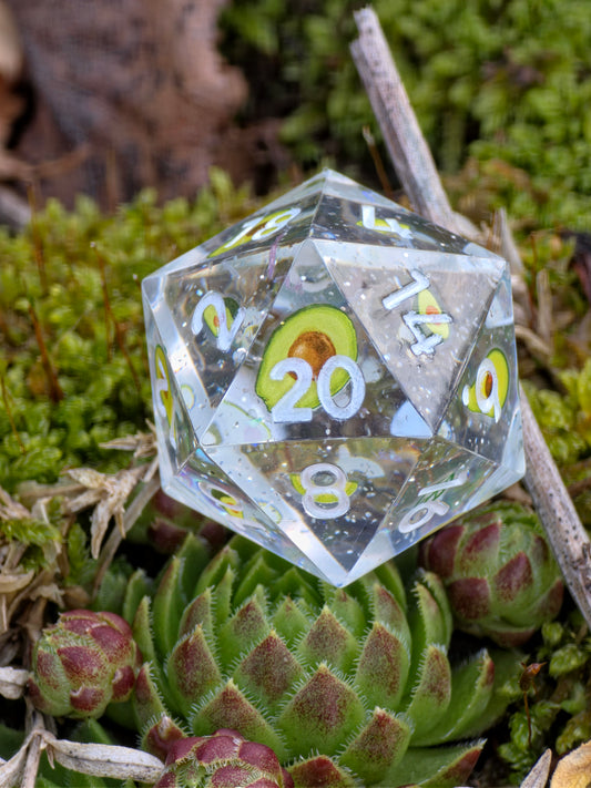 Handgemachter polyedrischer D20 „Avocado“ – transparenter Resinwürfel mit kleinen Avocado-Elementen und klarer Struktur.  
Handmade polyhedral D20 “Avocado” – transparent resin die with tiny avocado details and clear structure.