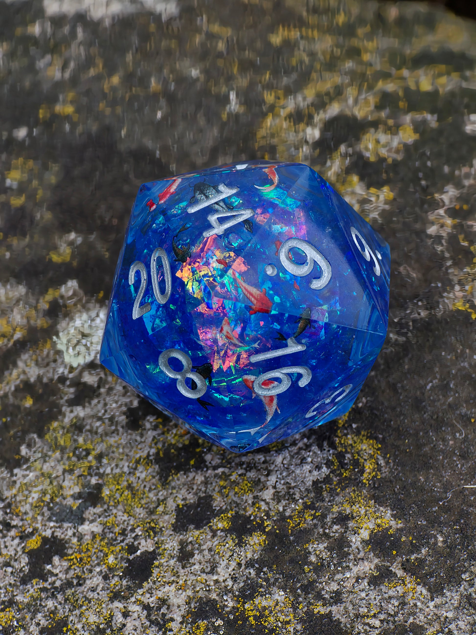 „Koi Pond“ D20 in der Hand – lebendige Farben, fließender Kern und leuchtende Fische im blauen Harz.  
“Koi Pond” D20 held in hand – vivid colors, flowing liquid core and glowing fish in blue resin.