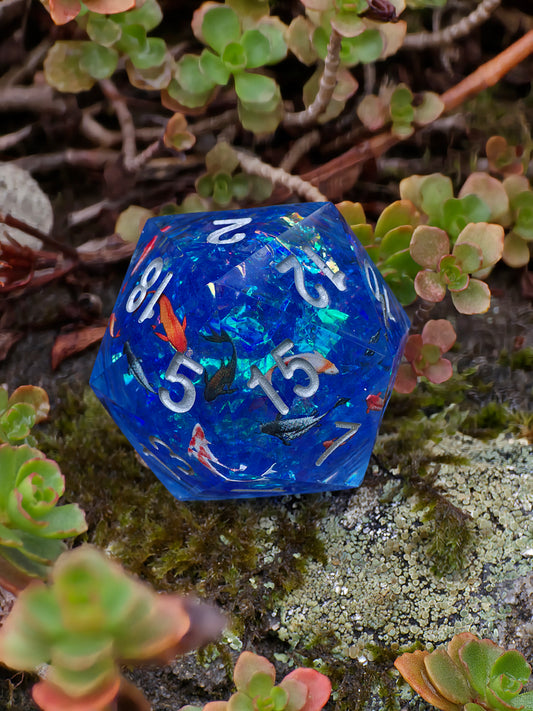 Handgemachter D20 „Koi Pond“ – großer Liquid Core Würfel mit schwimmenden Koi-Fischen in tiefblauem Harz.  
Handmade D20 “Koi Pond” – large liquid core die with floating koi fish in deep blue resin.